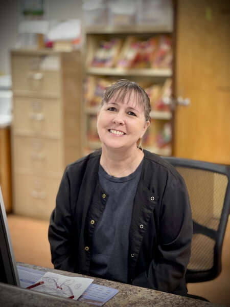 A smiling woman sits at a desk in an office environment, wearing a dark top and a black jacket.