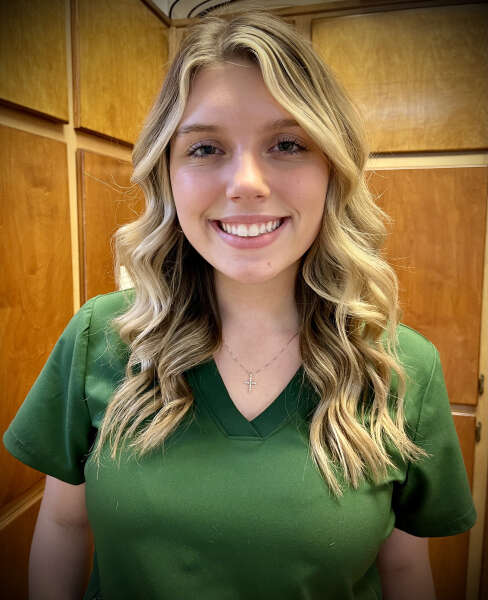 Smiling person with long, wavy blonde hair wearing a green shirt and a cross necklace.