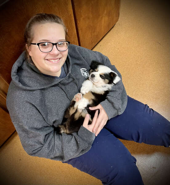 A person sitting on the floor, smiling and holding a small black and white puppy.