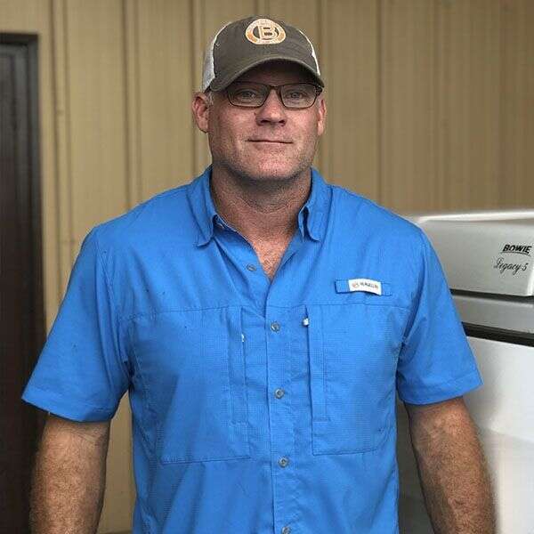 A man wearing a blue short-sleeve shirt, glasses, and a cap stands in front of a beige wall.