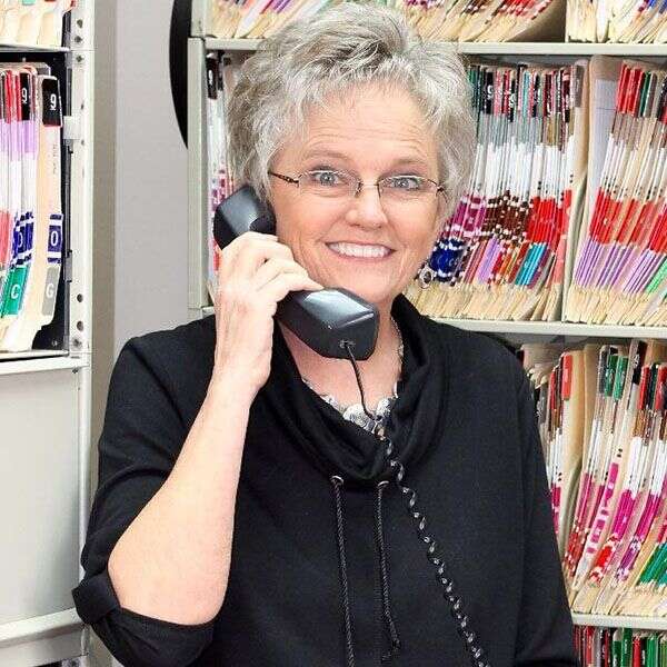A senior woman with short gray hair and glasses smiles while holding a phone, standing in front of shelves with files.