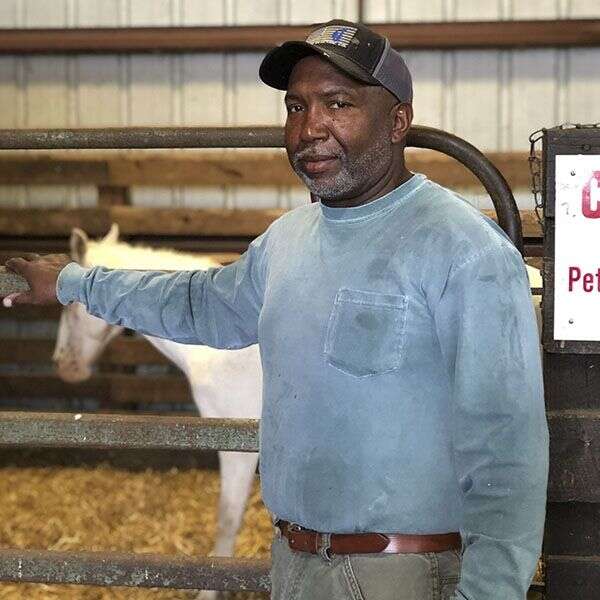A man in a blue shirt stands next to a horse in a stable, looking at the camera.