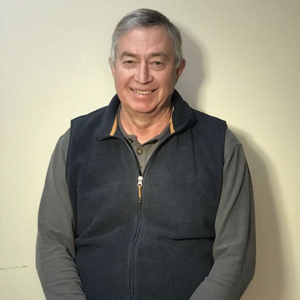 Older man smiling, wearing a dark vest over a long-sleeve shirt, standing against a plain background.